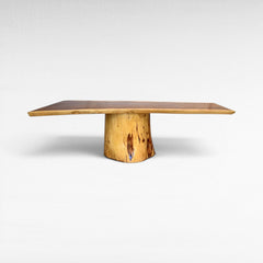 Chora Barefoot Luxury Living suar wood dining table with natural grain and live edge. Features a thick, solid wood slab top and a unique tree trunk pedestal base. Warm brown tones.